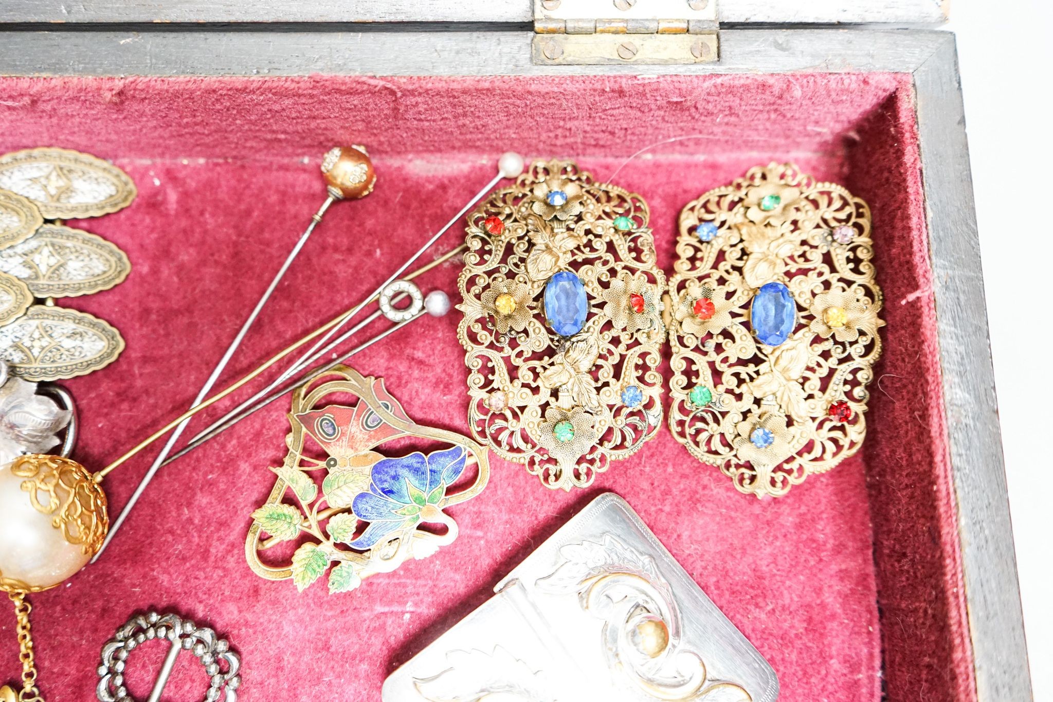 A group of belt buckles, clips and hatpins in a mahogany display case, 30.5cm
